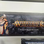 A Warhammer Age of Sigmar display sign featuring the game's logo, website, and an armored fantasy warrior holding a large axe.