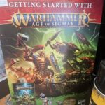 A Warhammer Age of Sigmar "Getting Started" box set featuring fantasy warriors battling on the cover, displayed on a store shelf next to a black and yellow spray can.