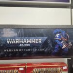 A store display sign for Warhammer 40,000 featuring a blue-armored Space Marine holding a gun, with the website warhammer40000.com shown below the logo.