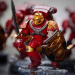 A close-up of a painted red armored miniature figurine holding a sword and shield, with similar figures blurred in the background.